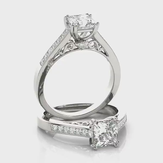 A silver engagement wedding ring set with a princess cut gem set on a filigree neck and channel set shank spinning on a viewing platform.