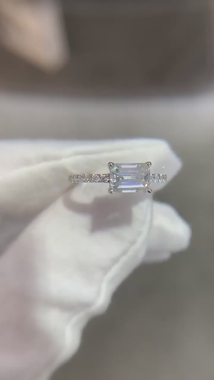 A silver East to West emerald cut moissanite ring with a pave band.