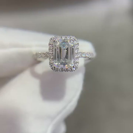 A hand displaying a silver emerald cut moissanite halo engagement ring.