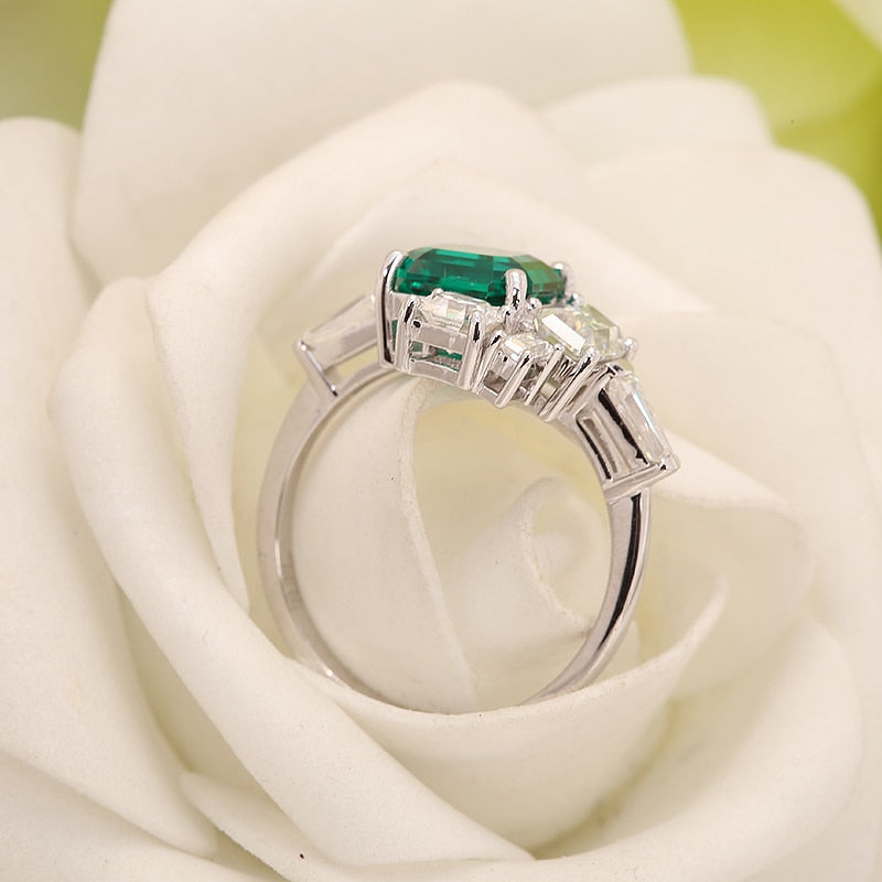 A silver asymmetrical ring set with a lab grown asscher cut emerald. To the right of the emerald is a moissanite baguette set east to west. To the left of the emerald is a emerald cut moissanite followed by a moissanite baguette set east to west. On top and slightly to the left of the emerald is a smaller asscher cut moissanite set next to a smaller emerald cut moissanite sitting atop of the larger emerald cut moissanite.