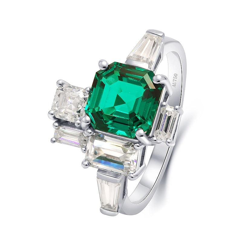 A silver asymmetrical ring set with a lab grown asscher cut emerald. To the right of the emerald is a moissanite baguette set east to west. To the left of the emerald is a emerald cut moissanite followed by a moissanite baguette set east to west. On top and slightly to the left of the emerald is a smaller asscher cut moissanite set next to a smaller emerald cut moissanite sitting atop of the larger emerald cut moissanite.