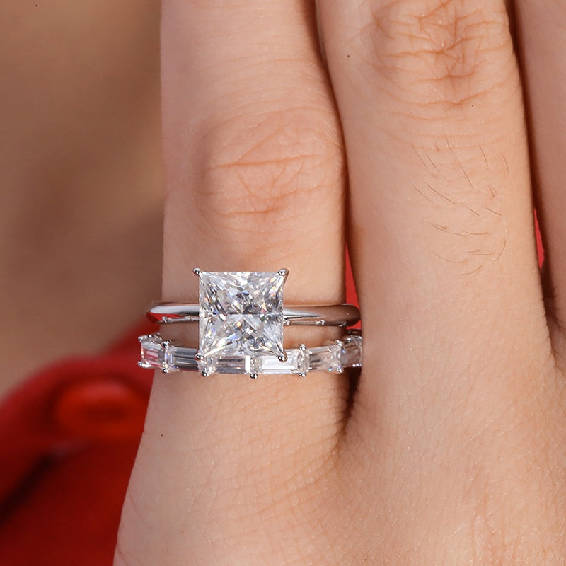 A silver ring set with a 2CT princess cut moissanite and paired with a moissanite baguette half eternity wedding band worn on a pinky finger.