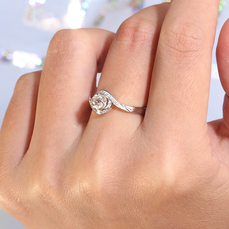 A hand wearing a silver round cut engagement ring set with a round moissanite on a pave bypass shank reminiscent of a wave.