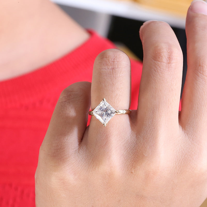 A hand wearing a gold solitaire band petal prong set with a 3CT princess cut moissanite, diagonally set with a tiny moissanite inset on each side of it.