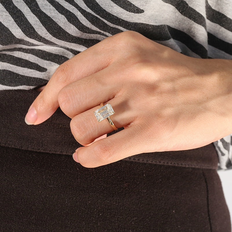 A hand wearing a gold solitaire ring basket set with a 4CT emerald cut moissanite.