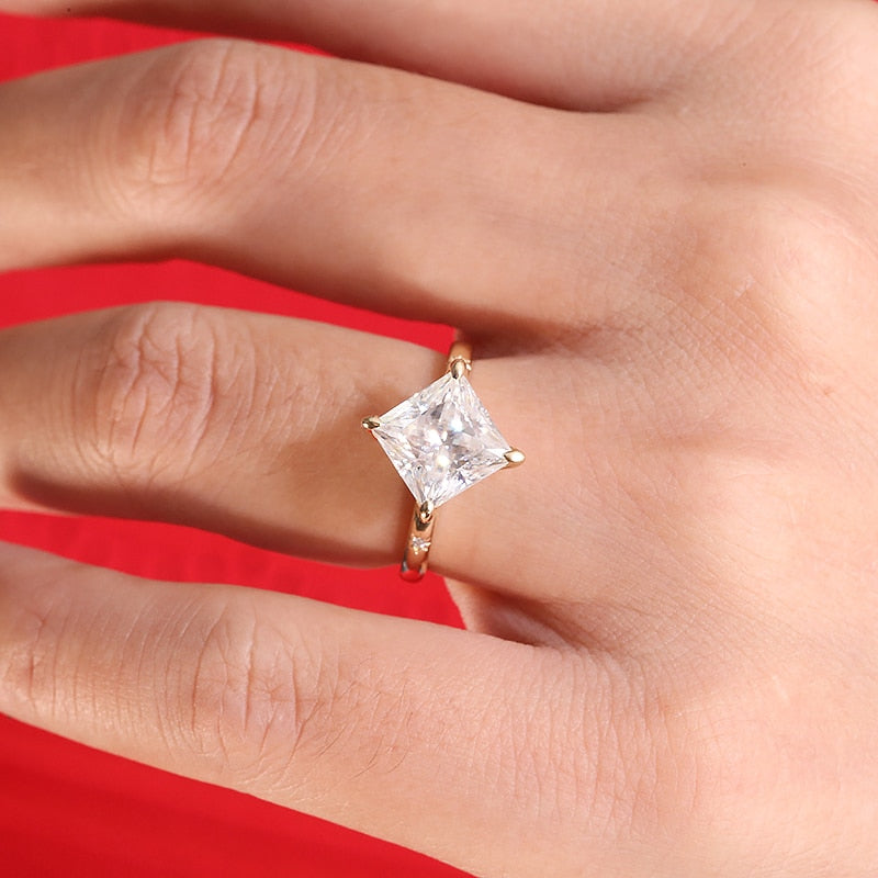 A hand wearing a gold solitaire band petal prong set with a 3CT princess cut moissanite, diagonally set with a tiny moissanite inset on each side of it.