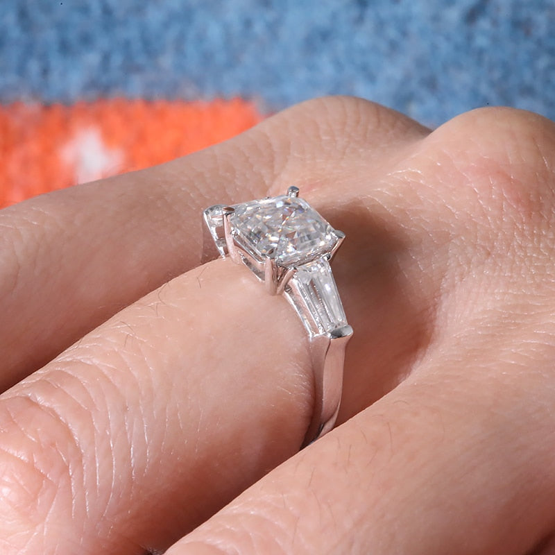 A hand wearing a silver 3 stone ring set with a Ascher cut moissanite nestled between two baguette moissanites.