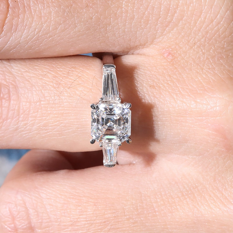 A hand wearing a silver 3 stone ring set with a Ascher cut moissanite nestled between two baguette moissanites.