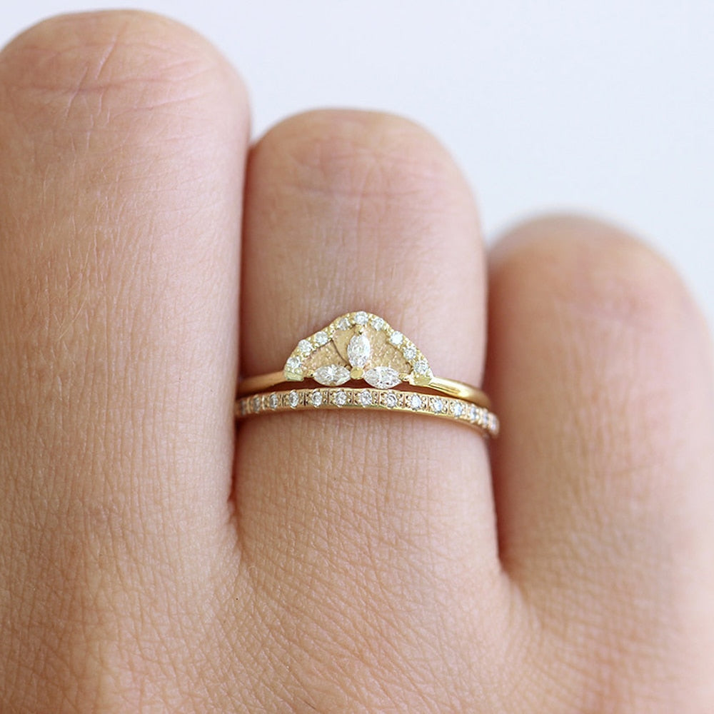 A hand wearing a gold crown engagement ring set with 3 small marquise cut moissanites and lined in small round moissanites accompanied by a matching wedding ring.