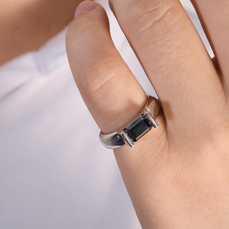 A hand wearing a silver ring with an emerald cut black moissanite side east to west in a tension setting.
