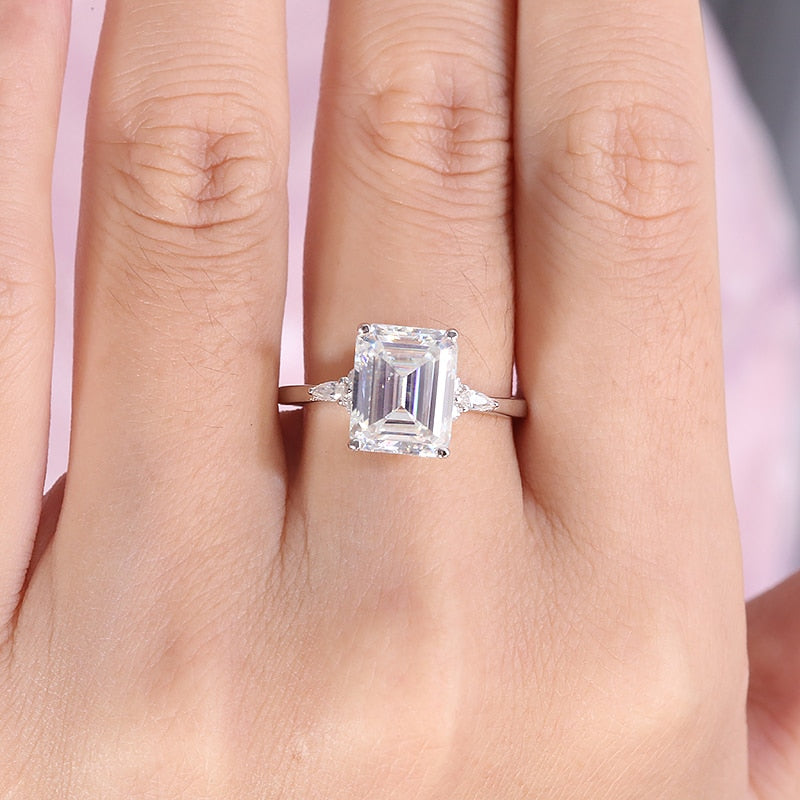 A hand wearing a silver ring with a 5CT emerald cut moissanite set between small round and small pear cut moissanites.