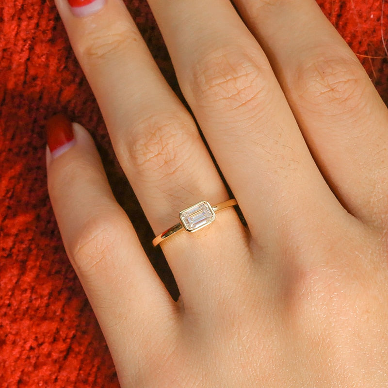 A hand wearing a silver ring that has a emerald cut moissanite bezel set east to west.