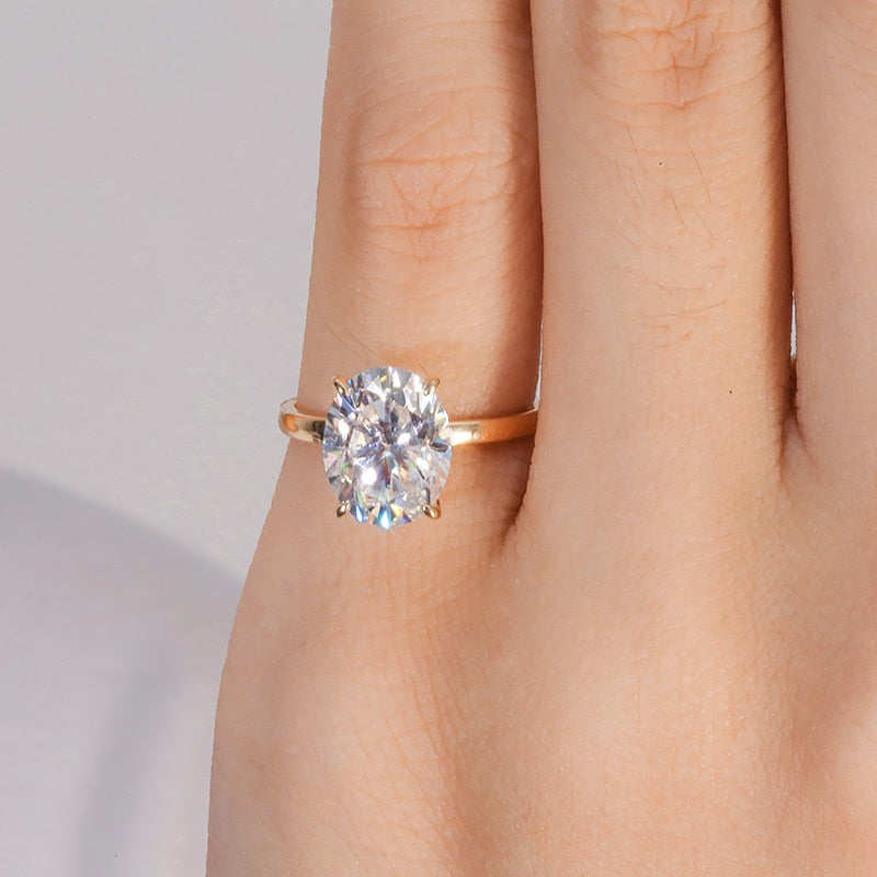 A hand wearing a gold band solitaire set with petal style prongs holding a 3CT oval moissanite.