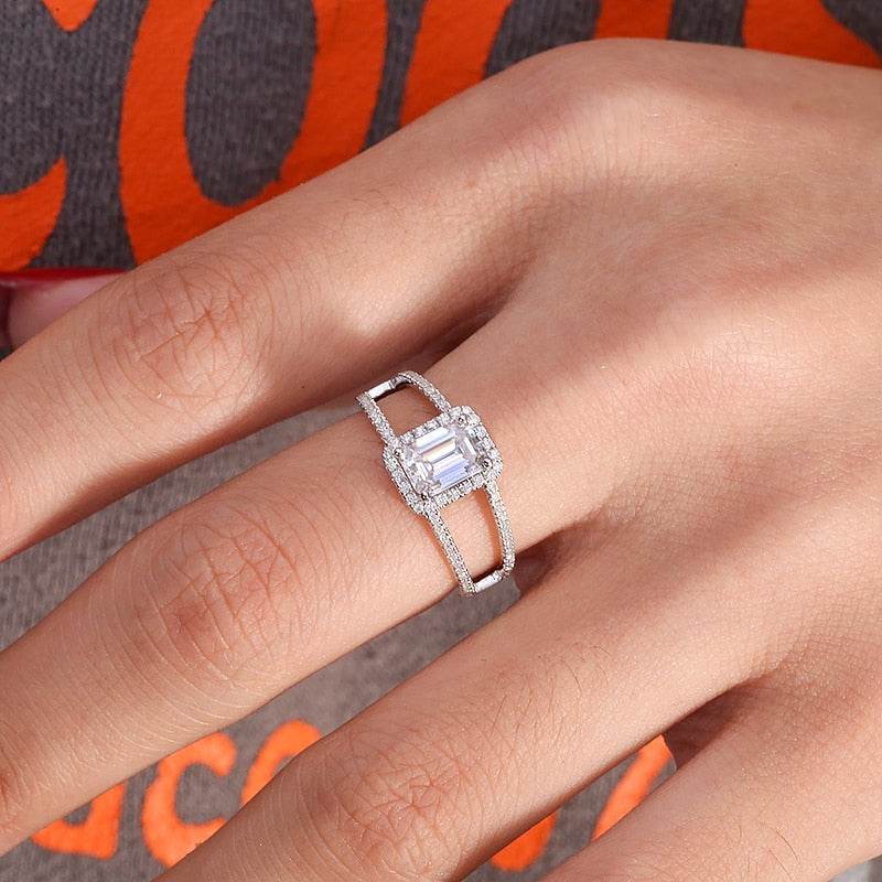 A hand wearing a silver ring set with a emerald cut moissanite with a wide split pave band.