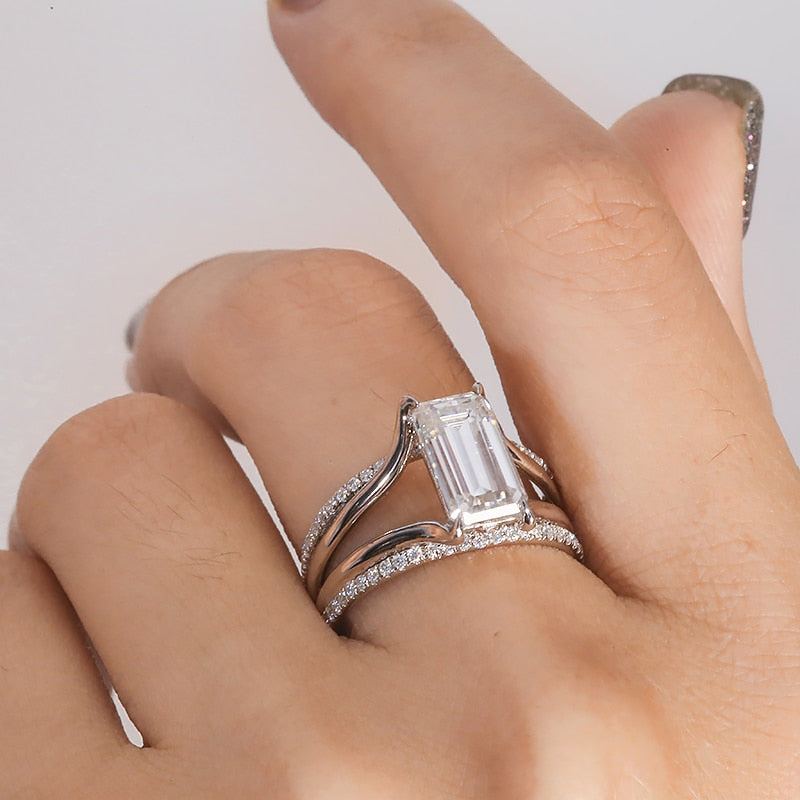 A hand wearing a silver split shank ring set with a 3CT emerald cut moissanite and a row of pave gems along the top and bottom band.