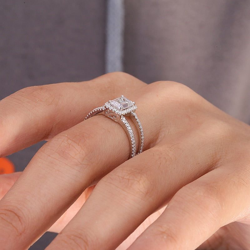A hand wearing a silver ring set with a emerald cut moissanite with a wide split pave band.