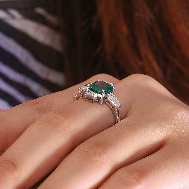 A hand wearing a silver asymmetrical ring set with a lab grown asscher cut emerald. To the right of the emerald is a moissanite baguette set east to west. To the left of the emerald is a emerald cut moissanite followed by a moissanite baguette set east to west. On top and slightly to the left of the emerald is a smaller asscher cut moissanite set next to a smaller emerald cut moissanite sitting atop of the larger emerald cut moissanite.