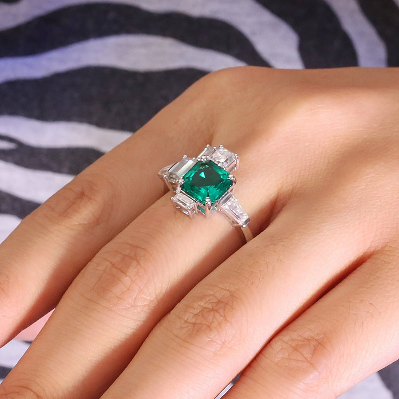 A hand wearing a silver asymmetrical ring set with a lab grown asscher cut emerald. To the right of the emerald is a moissanite baguette set east to west. To the left of the emerald is a emerald cut moissanite followed by a moissanite baguette set east to west. On top and slightly to the left of the emerald is a smaller asscher cut moissanite set next to a smaller emerald cut moissanite sitting atop of the larger emerald cut moissanite.