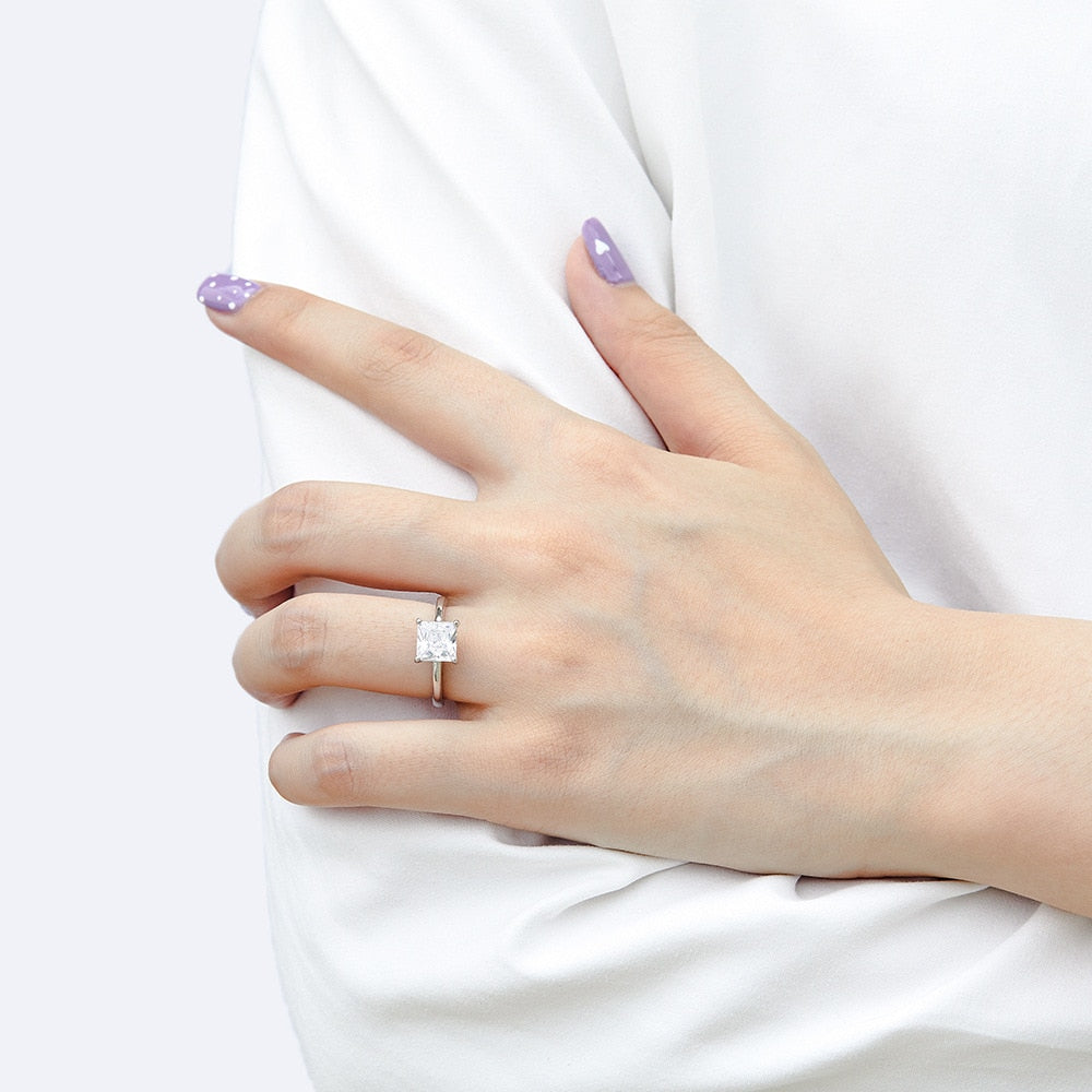 A hand wearing a silver ring set with a princess moissanite.