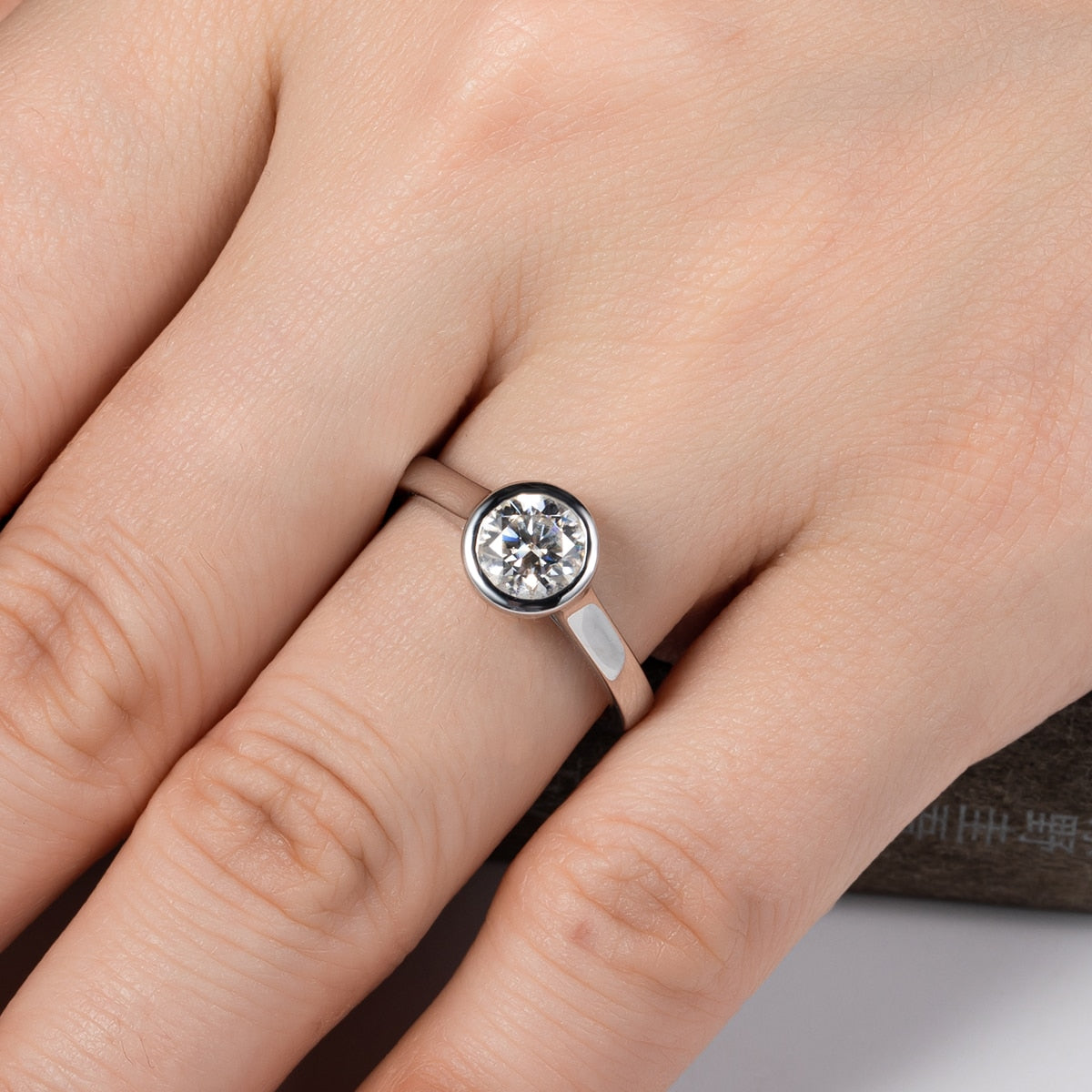 A hand modeling a sterling silver engagement ring with a 1CT round cut moissanite in a bezel setting.
