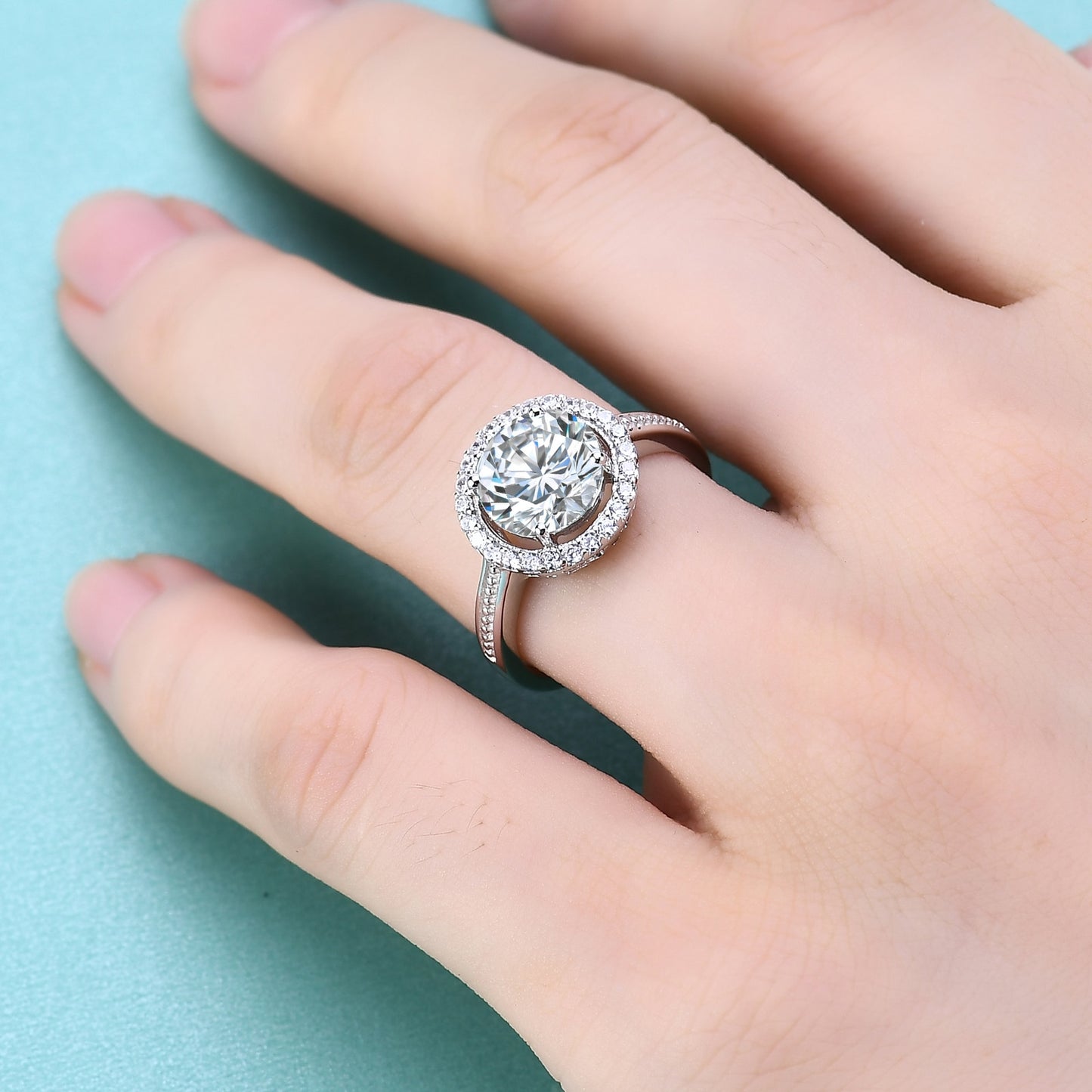A silver halo ring set with a 2CT round moissanite encircled by a clear gem halo and set on a textured band worn on a ring finger.