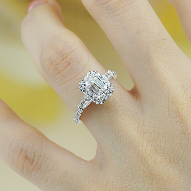 A hand wearing a silver engagement ring set with an emerald cut moissanite and is a hybrid 3 stone halo style.