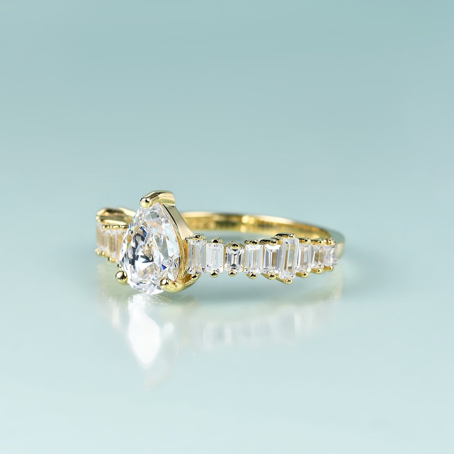 A gold ring with a tear drop shape moissanite on a band of varying side emerald cut gems set horizontally.
