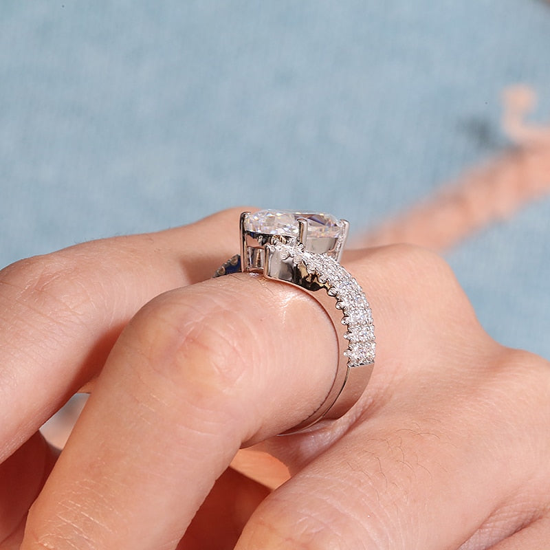 A hand wearing a silver ring set with a oval 6CT moissanite set in a triple row half pave bypass style band.