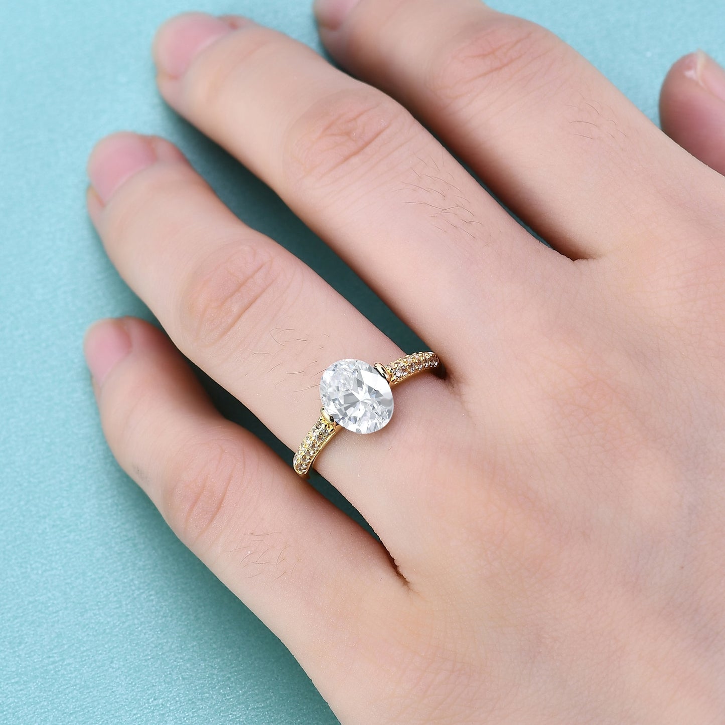 A gold tension set ring with a oval moissanite and double row pave band worn on the ring finger of a hand.