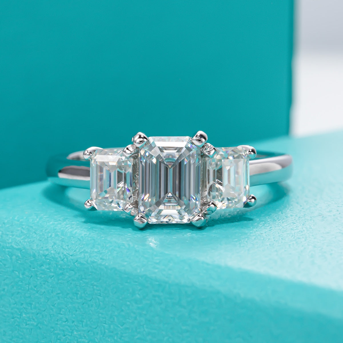 A silver 3 stone ring with a 1CT emerald cut moissanite set between two smaller emerald cut moissanites.