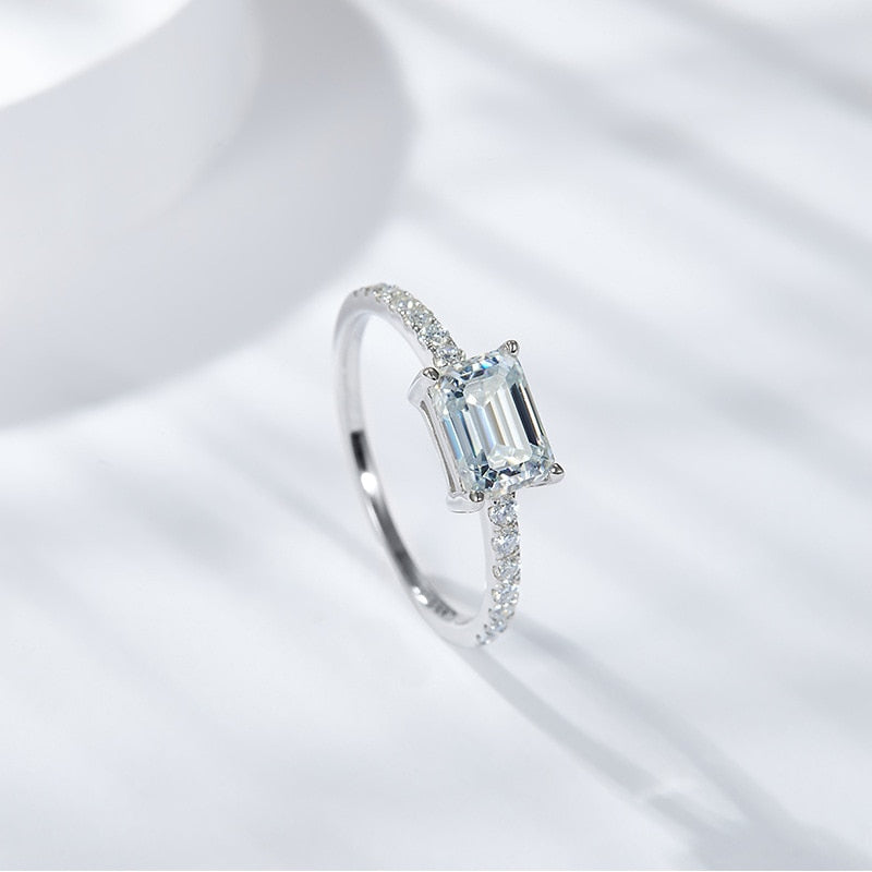 A silver East to West emerald cut moissanite ring with a pave band.