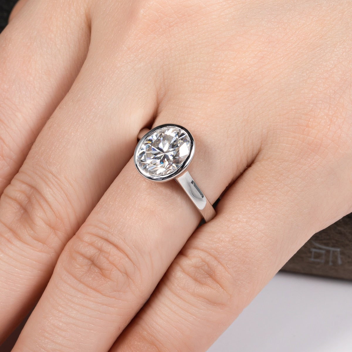 A hand wearing a 3CT oval cut moissanite set in a sterling silver bezel style setting.