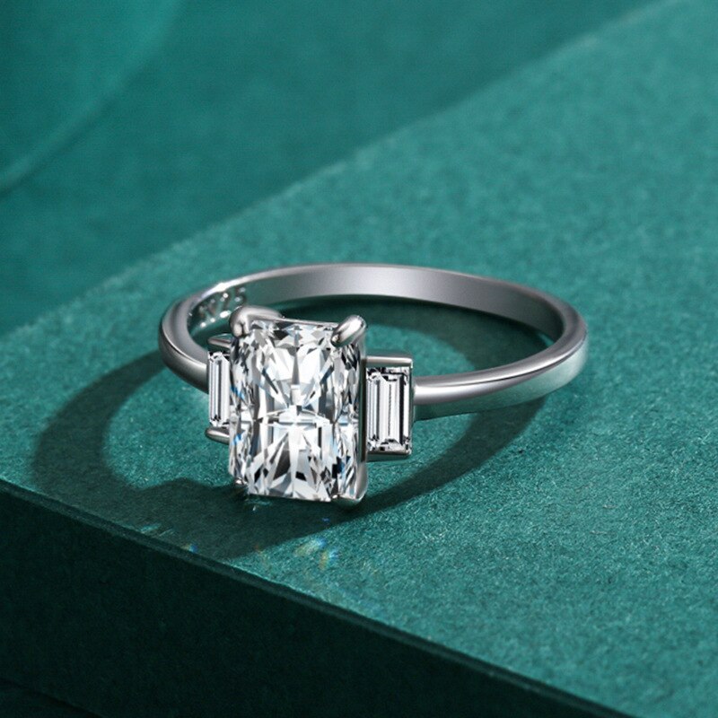 A silver ring featuring a 3CT radiant cut moissnite set between two smaller emerald cut gems.