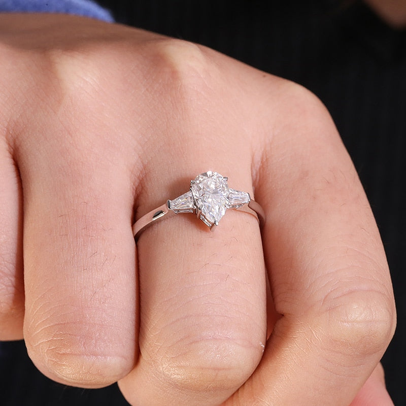 A hand wearing a silver 3 stone ring set with a 1.5CT pear cut moissanite set between two smaller baguettes.