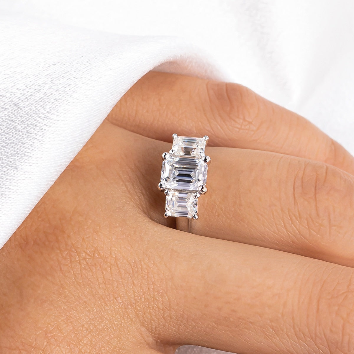 A hand wearing silver 3 stone ring with a 1CT emerald cut moissanite set between two smaller emerald cut moissanites.
