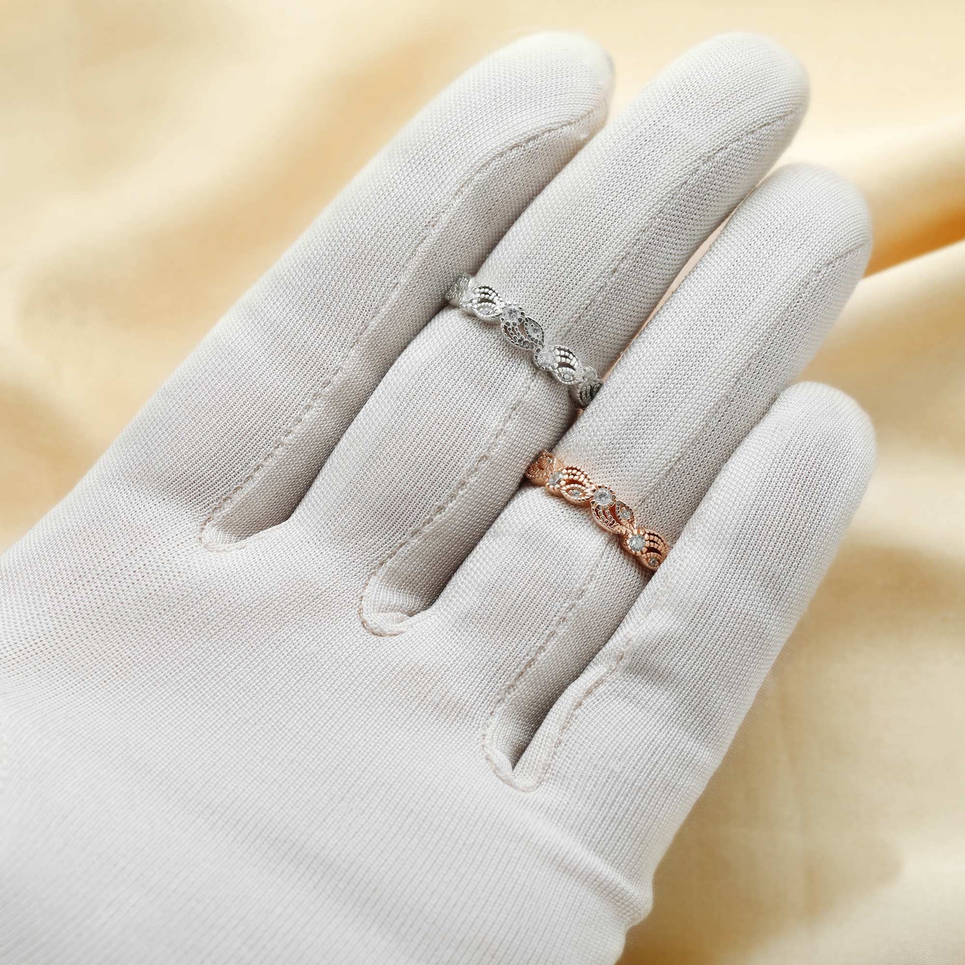 A hand wearing one silver and one rose gold swirl and leaf designed bands set with clear small gems.