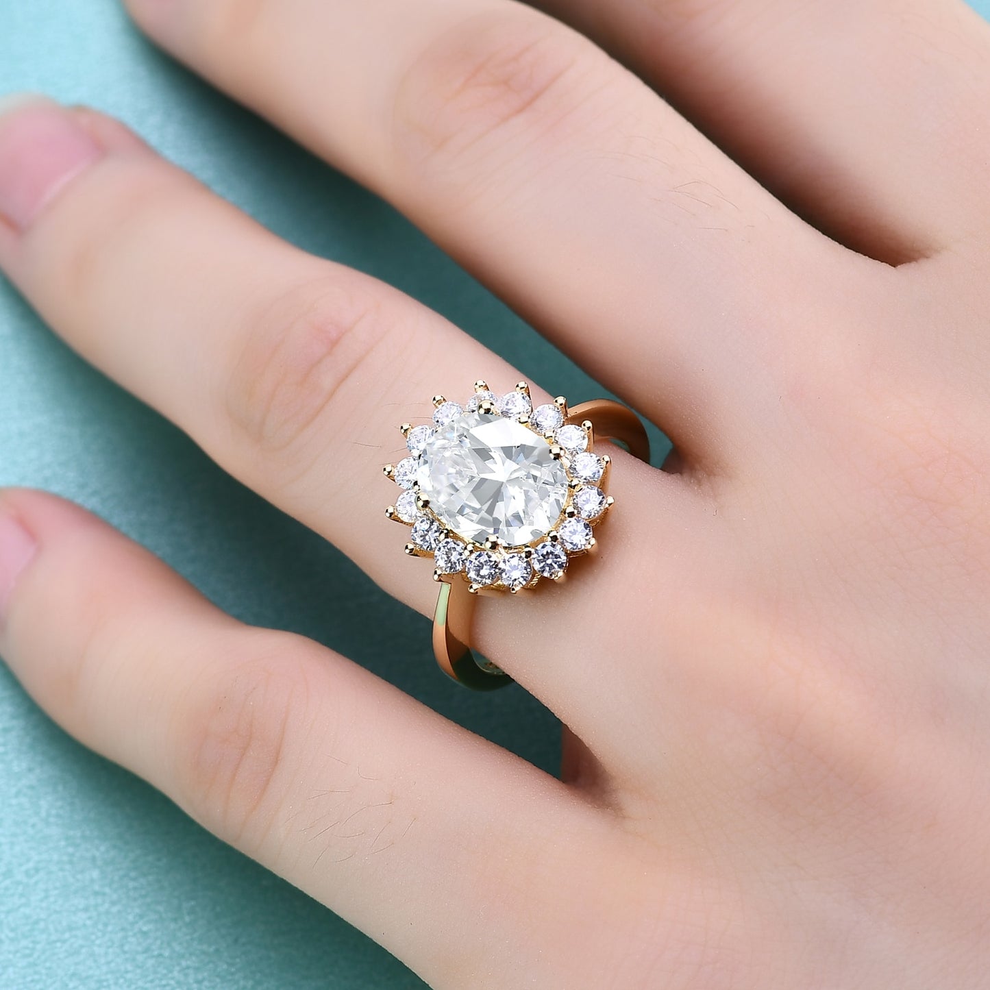 A gold halo ring set with a 3CT oval cut moissanite set in the middle of a ring of clear gems, worn on a ring finger.