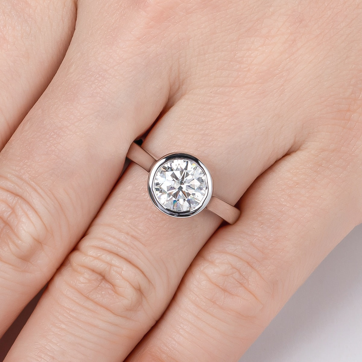 A hand wearing a sterling silver engagement ring with a 3CT round cut moissanite in a bezel setting.