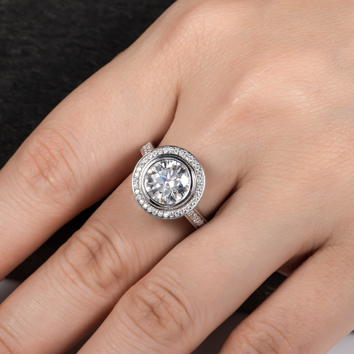 A vintage style ring with 3CT moissanite bezel set surrounded with a halo and pave band worn on a hand.