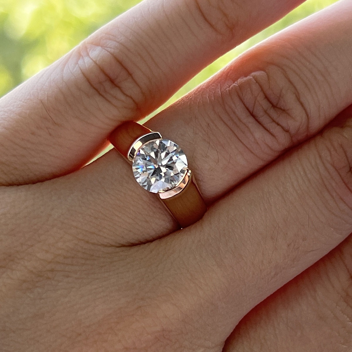 A rose gold plated round cut moissanite tension set in a floating half bezel setting on a ring finger.