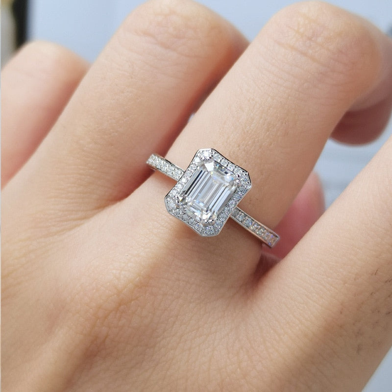 A hand wearing a silver halo ring set with an emerald cut moissanite.