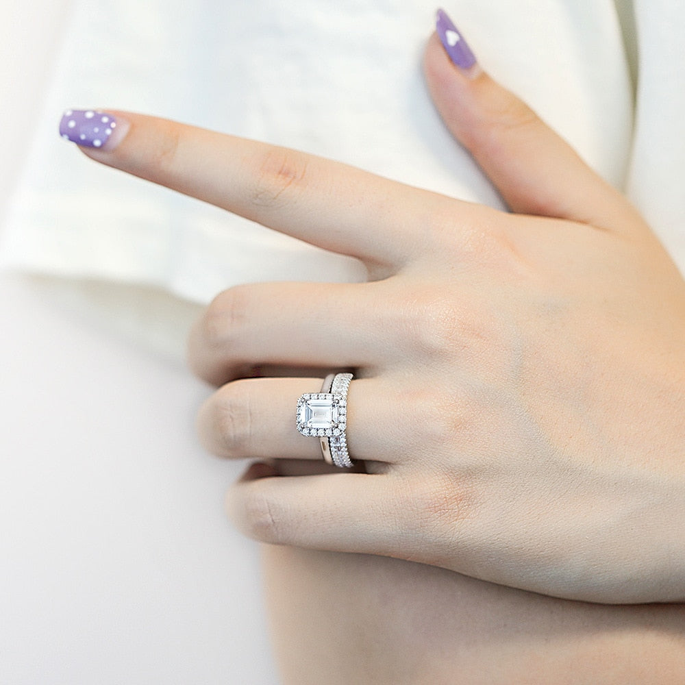 A hand wearing a silver engagement and wedding set that includes a emerald cut halo ring and a matching round and emerald cut half eternity ring.
