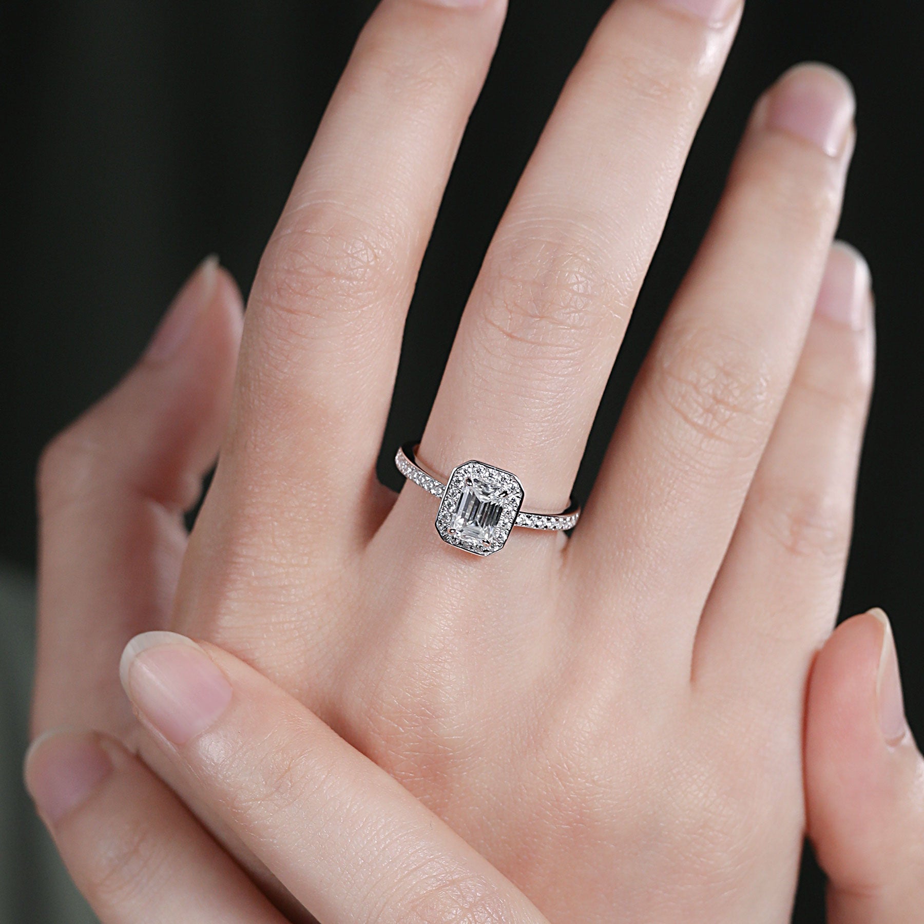 A hand wearing a silver emerald cut halo ring.