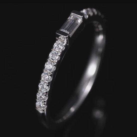 A silver stackable wedding ring tension set with a small emerald cut gem on a pave band spinning on a viewing platform.